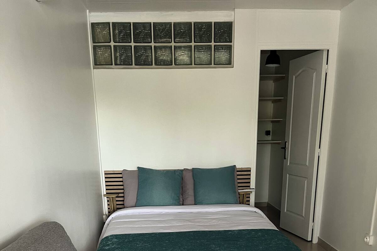 A bedroom with a bed, a grey armchair, a white wall with a window, and a door leading to a closet.