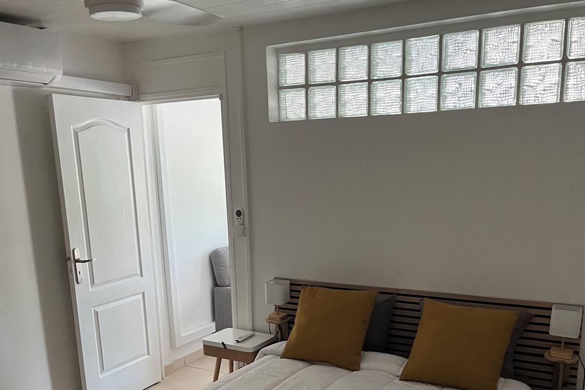 A bedroom with a bed, a ceiling fan, a window with blinds, a bedside table with a lamp, and a door.