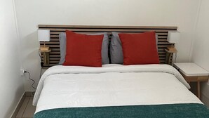 A neatly made bed with a white and teal bedspread, two red and gray pillows, and wooden headboard with bedside lamps. - Résidence hôtelière Le Manguier Panoramique (Trois-Ilets)