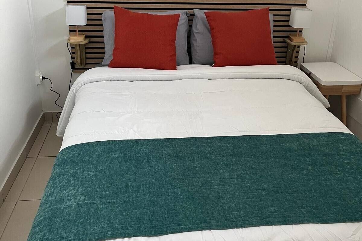 A neatly made bed with a white and teal bedspread, two red and gray pillows, and wooden headboard with bedside lamps.