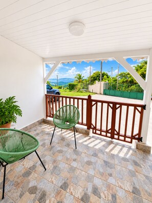A covered outdoor area with a stone floor, a green chair, a potted plant, and a view of a blue car and greenery. - Résidence hôtelière Le Manguier Panoramique (Trois-Ilets)