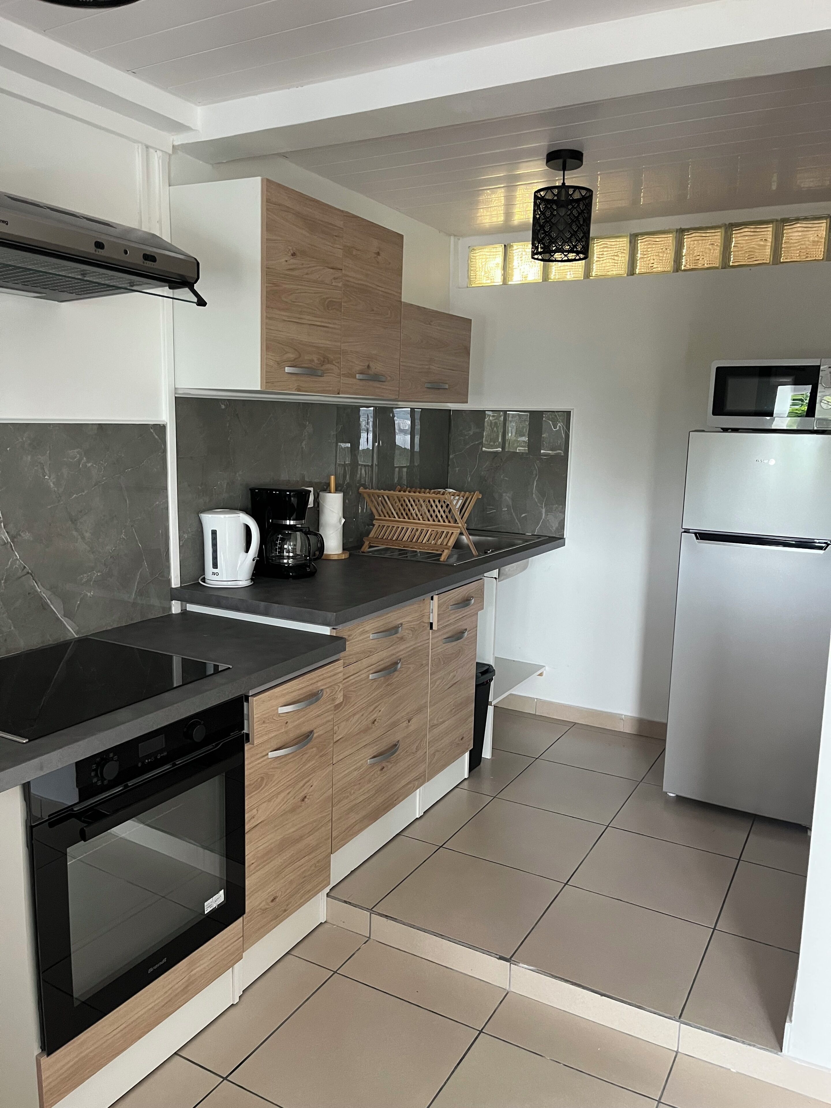 Una cocina moderna con placa de inducción negra, armarios de madera, refrigerador de acero inoxidable y lámpara colgante negra.