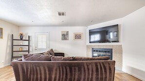 A living room with a brown sofa, a flat-screen TV mounted above a fireplace, and a wooden floor. - Gated home w/ backyard retreat (Sacramento)