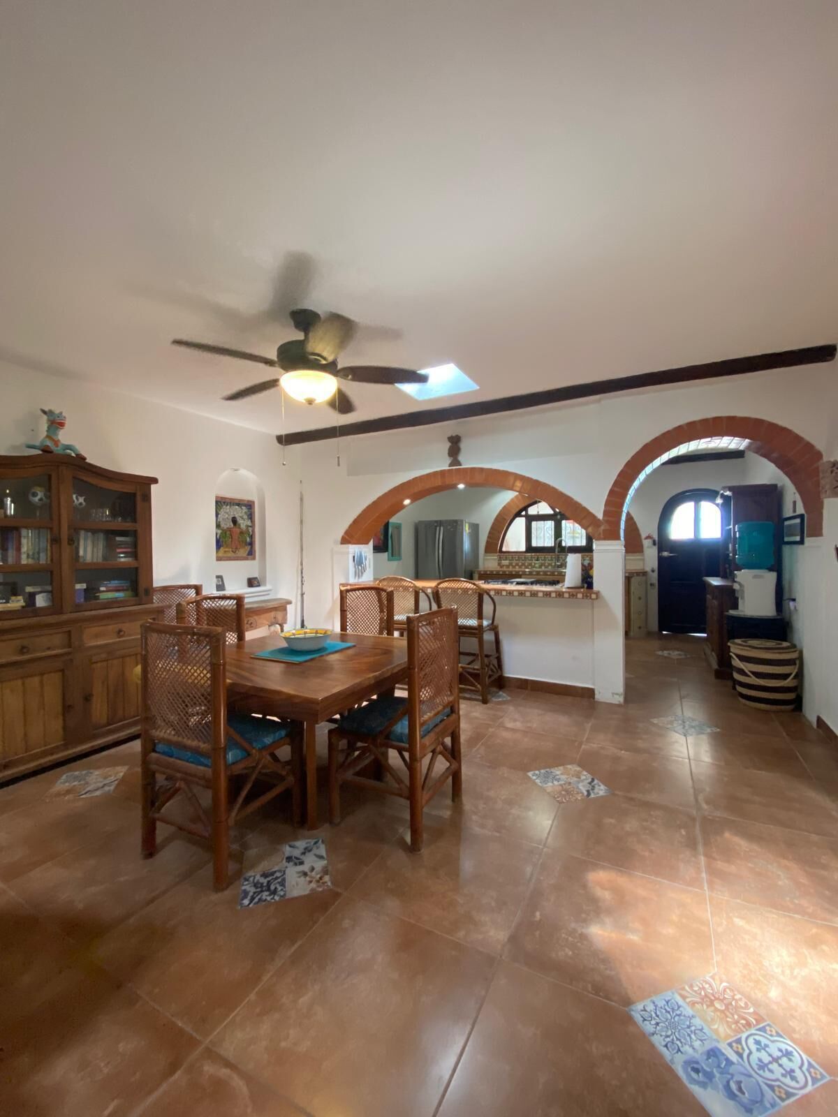 A dining area with a wooden table and chairs, a ceiling fan, and a kitchen in the background.
