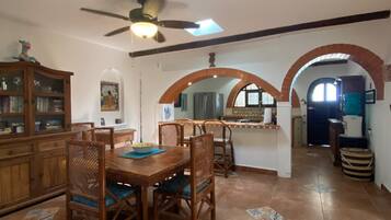 A dining area with a wooden table and chairs, a ceiling fan, and a kitchen in the background.