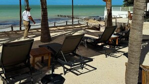 Ein Sitzbereich am Strand mit Liegestühlen und einem Tisch, Palmen und Blick aufs Meer.