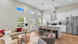 A spacious living area with a sofa, a coffee table, and a dining table. There is a kitchen with white cabinets and stainless steel appliances.