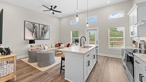 A spacious living area with a sofa, ottoman, and a dining table. There is a kitchen with a large island, cabinets, and a window.