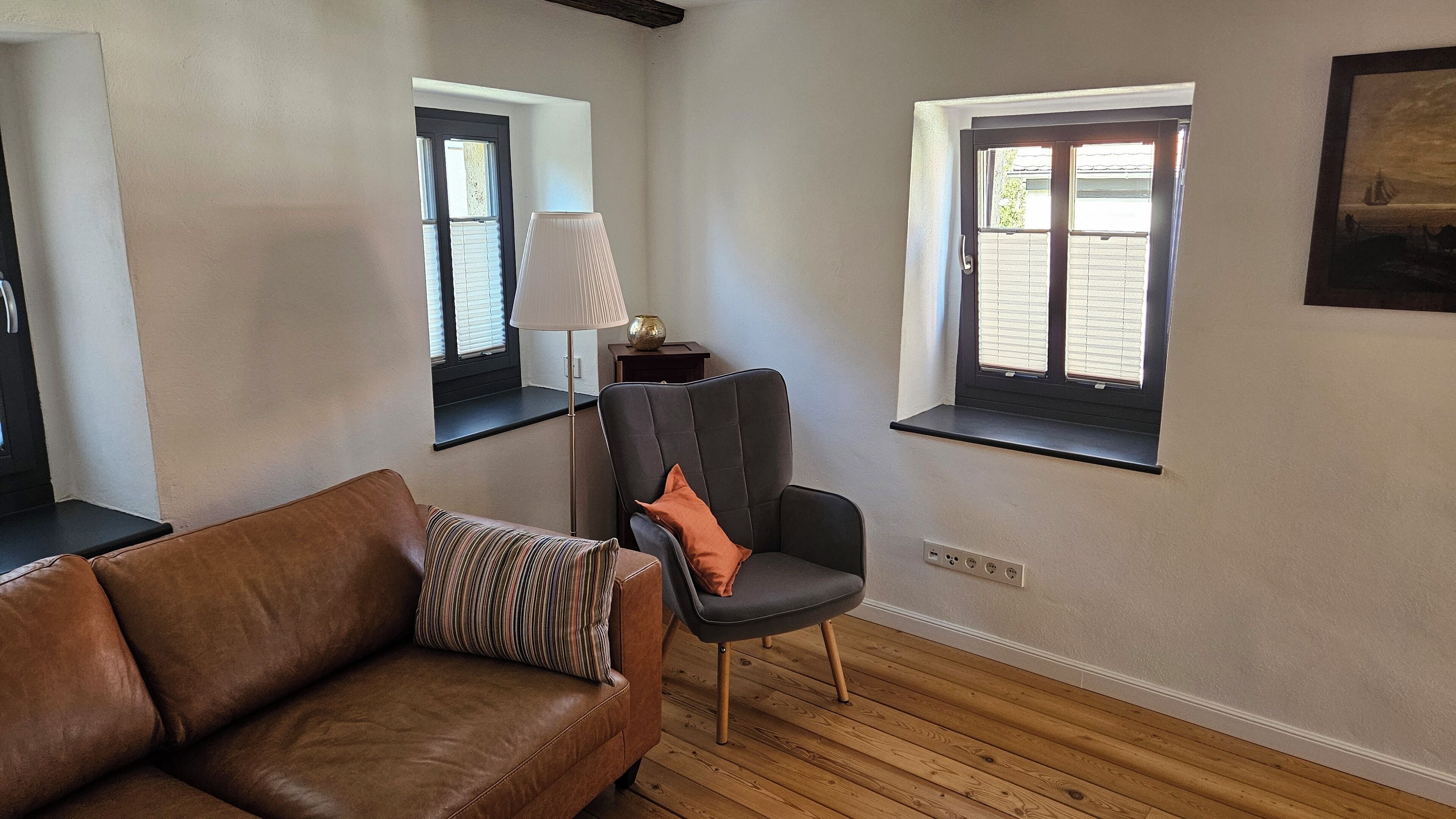 A living room with a brown leather sofa, a black chair with an orange cushion, a floor lamp, and two windows with blinds.