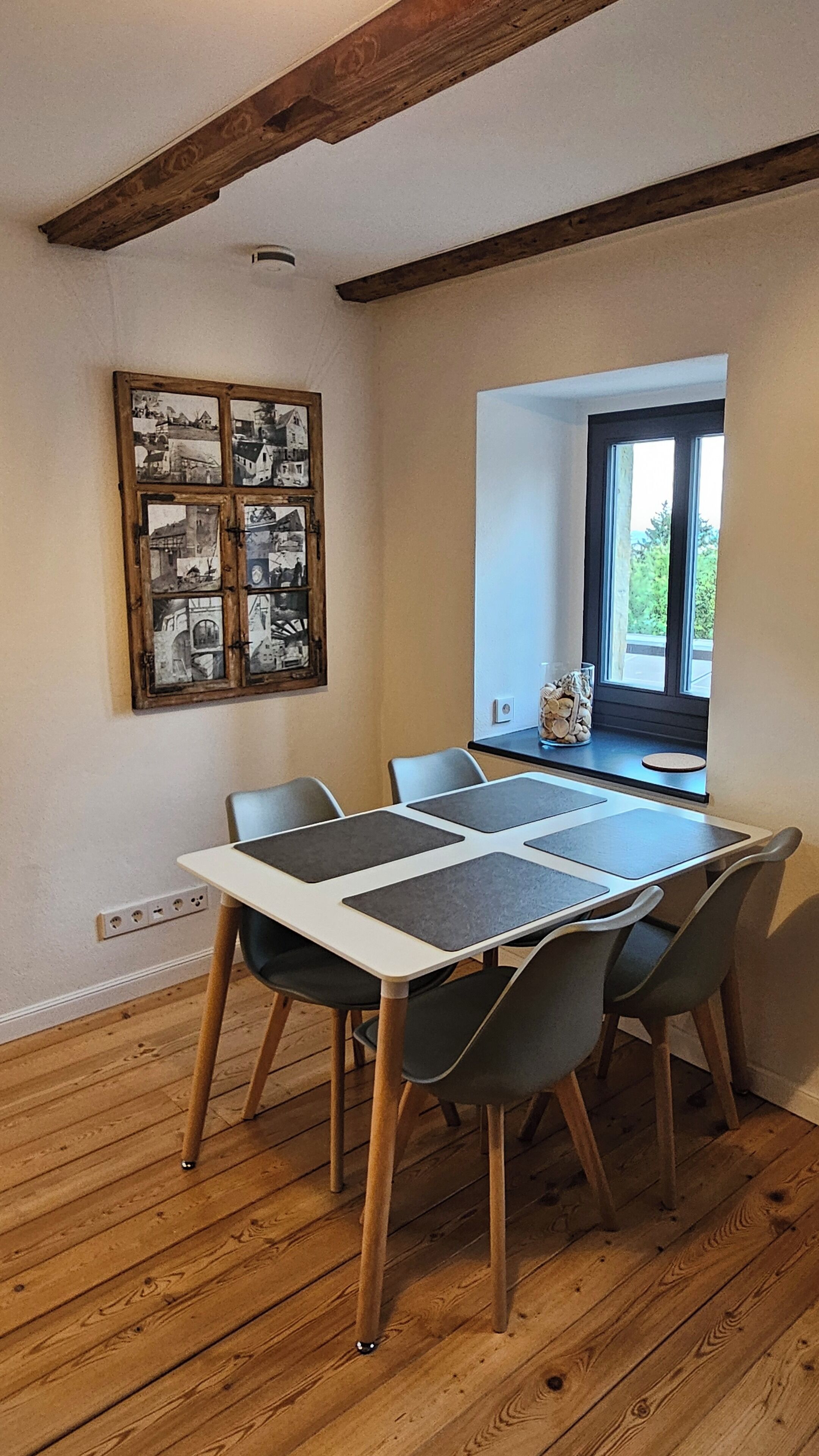 Un comedor con una mesa y sillas de madera, un collage enmarcado en la pared, y una ventana con vistas a zonas verdes.