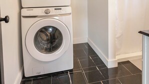 Uma lavanderia com duas máquinas de lavar Samsung brancas, chão de azulejos pretos e um chuveiro com cortina branca.