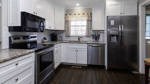 Una cocina moderna con armarios blancos, electrodomésticos de acero inoxidable, un ventanal con cortina y suelo de madera oscura.