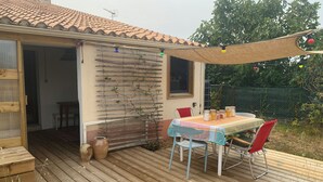 A wooden deck with a table and chairs, a pergola with string lights, and a view of a garden. - Comfortable tiny house with kitchen and summer bathroom by the sea (Brem-sur-Mer)