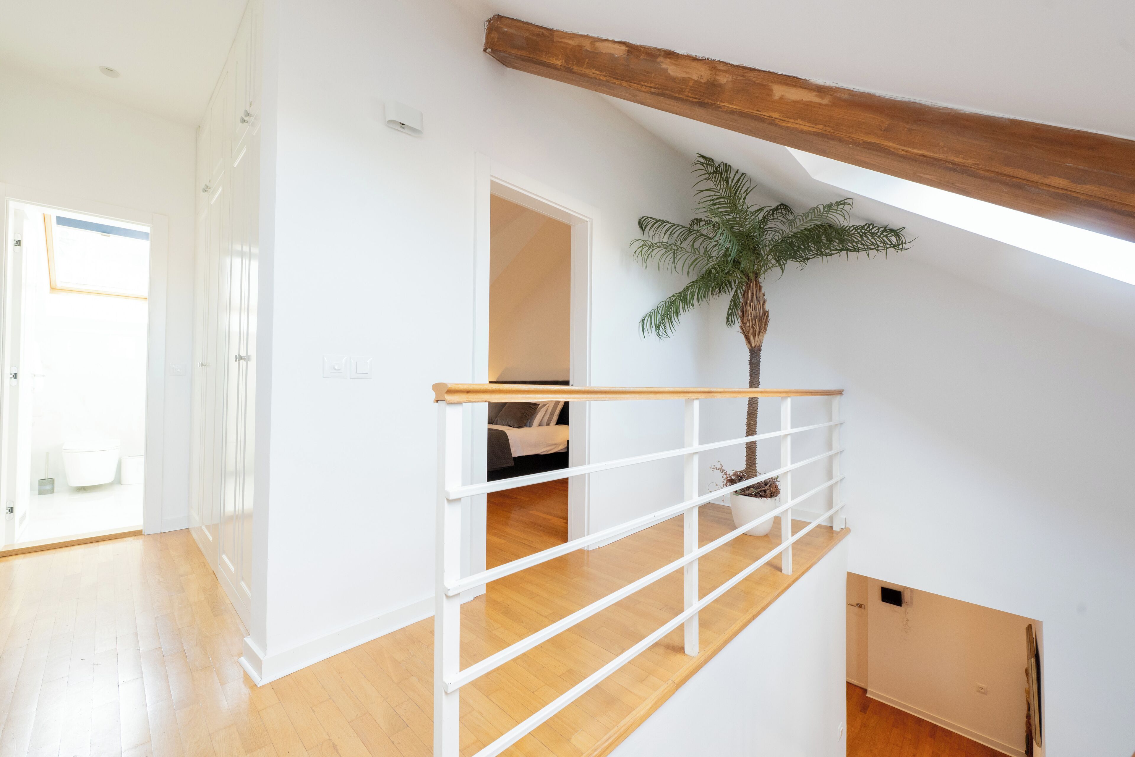 Une pièce spacieuse dotée d’un plancher en bois, de murs blancs et d’un plafond avec poutres apparentes. Un escalier avec une rampe blanche mène à un niveau supérieur.