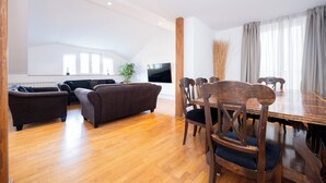 Un vaste espace de vie comprenant une table à manger en bois, des canapés foncés et un téléviseur à écran plat fixé au mur.