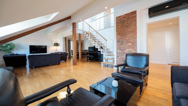 Un vaste espace de vie doté d’un plancher en bois, d’un canapé en cuir noir, d’un fauteuil noir et d’une table basse. Un escalier avec une rampe en verre mène à un niveau supérieur.