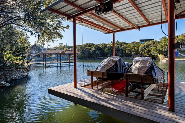 A wooden dock with covered seating and a canopy, overlooking a calm lake with a dock and house in the background.