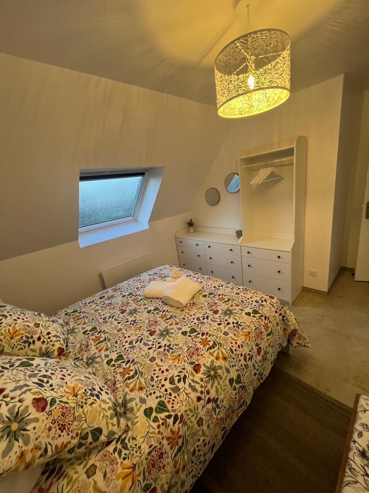 Une chambre à coucher avec un lit, une commode, une petite fenêtre et un luminaire suspendu.