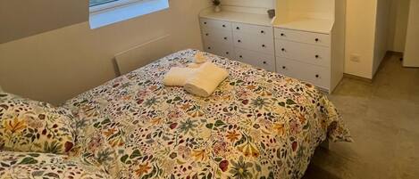 Une chambre à coucher avec un lit, une commode, une petite fenêtre et un luminaire suspendu.