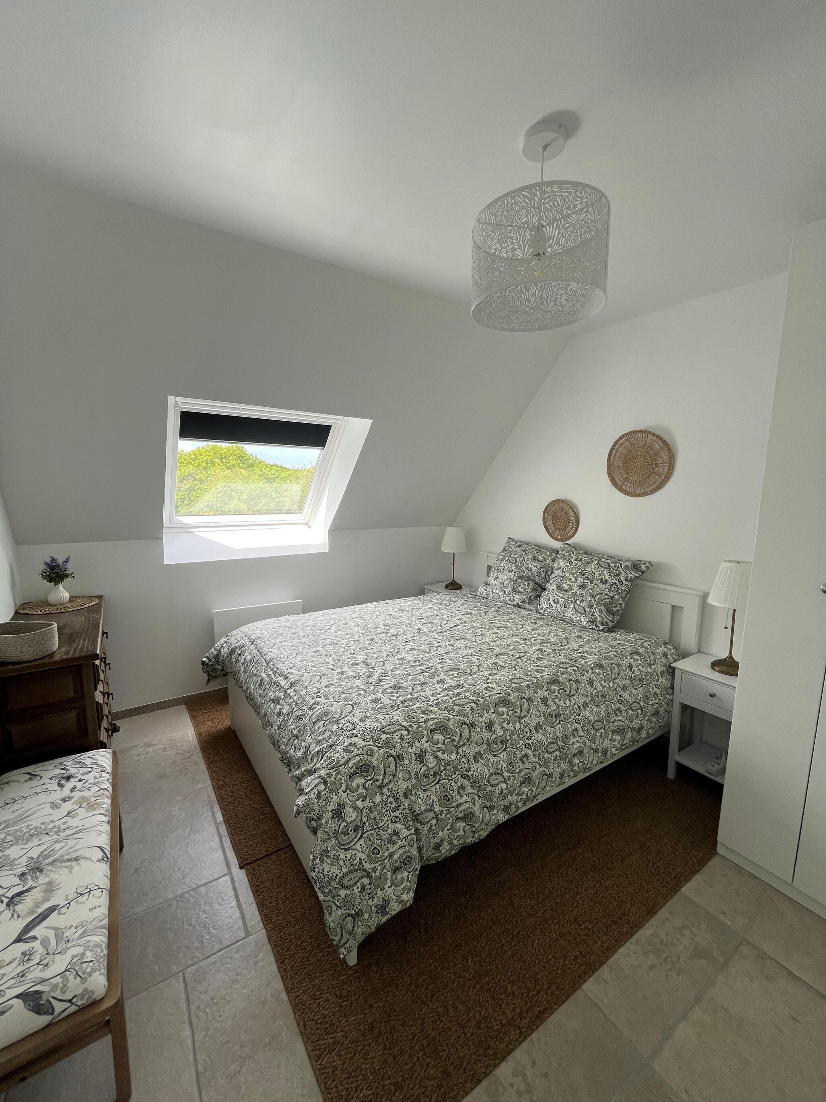 Une chambre à coucher comprenant un lit, une fenêtre, une table de chevet, une lampe, une commode, une chaise et un tapis.