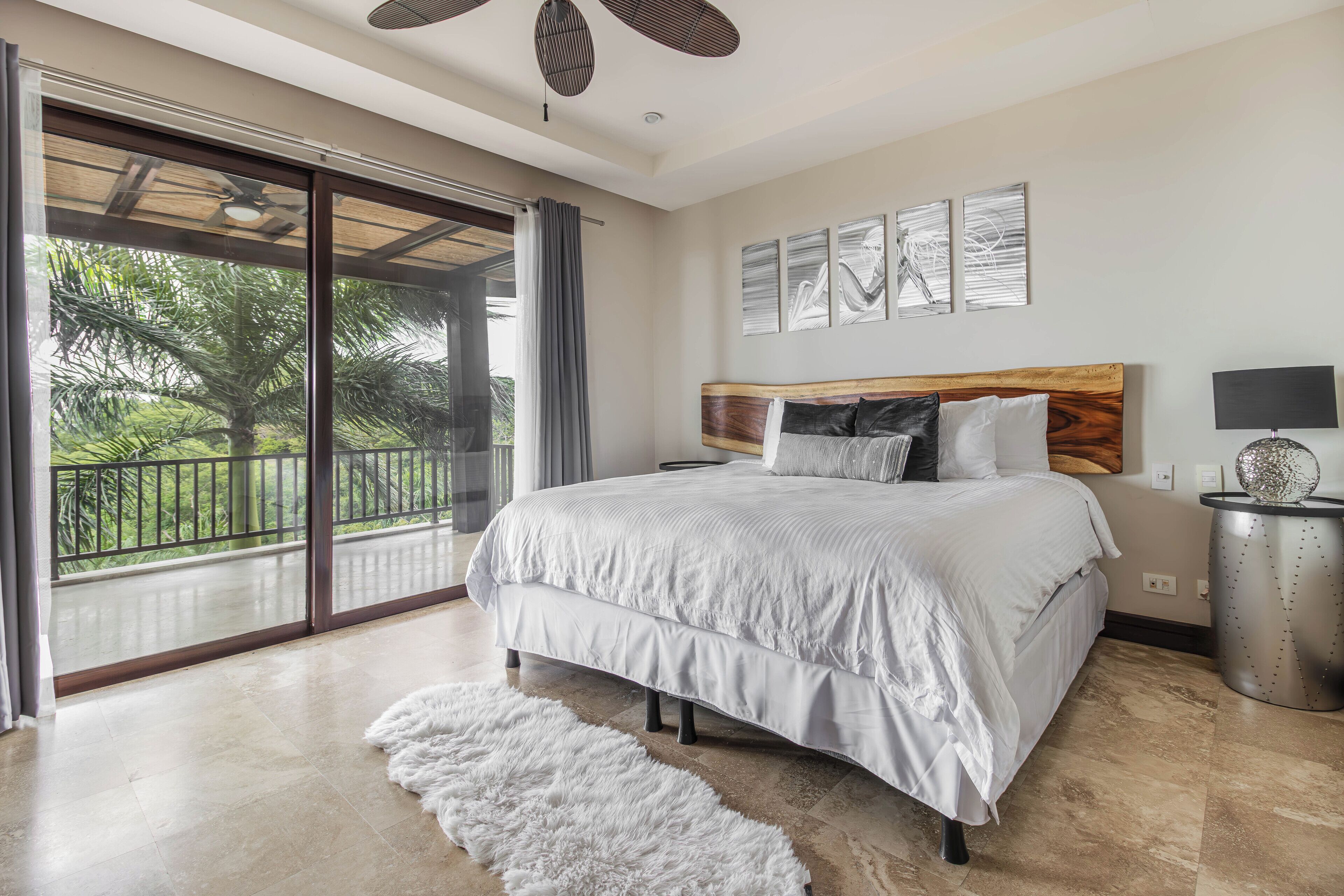 A bedroom with a large bed, a view of greenery through a sliding door, and a ceiling fan.