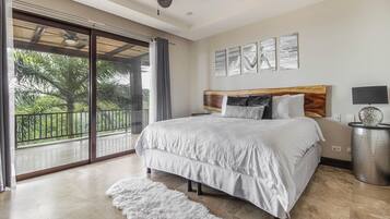 A bedroom with a large bed, a view of greenery through a sliding door, and a ceiling fan.