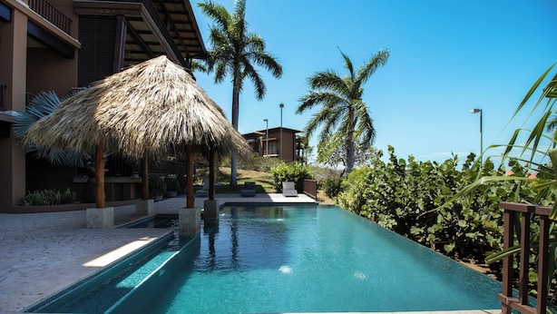 A swimming pool with a thatched umbrella, palm trees, and a building with a balcony.