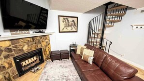 A living room with a stone fireplace, a brown leather sofa, a mounted TV, and a staircase.