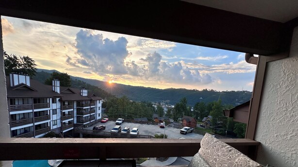 A balcony with a view of a parking lot and buildings, a dining table with a plate, and a patterned cushion.