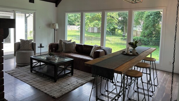 Un salón espacioso con techo alto, ventanas grandes, una mesa de comedor de madera con taburetes tipo bar, un sofá de cuero y una mesa de centro de vidrio.