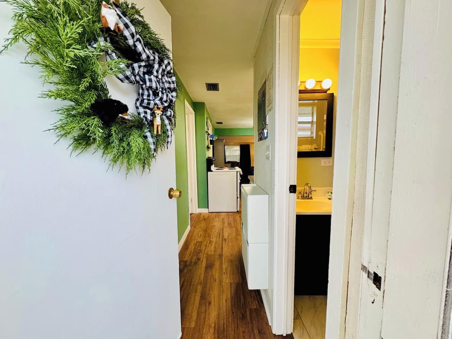 A hallway with a green wall, wooden flooring, a white door, and a small kitchen area with a sink and countertop visible.