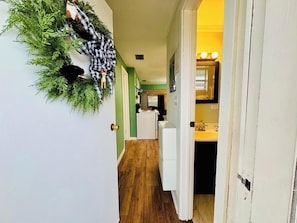 Un pasillo con pared verde, suelo de madera, una puerta blanca y una pequeña zona de cocina con un lavabo y encimera a la vista.