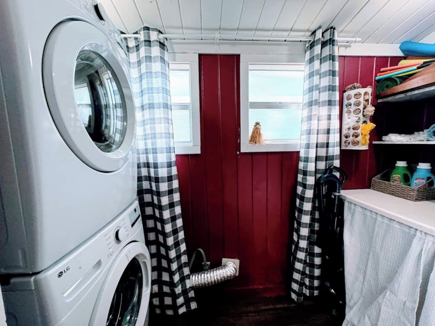 Een wasserette met een voorlader wasmachine en droger, geruite gordijnen, een klein raam en een plank met schoonmaakspullen.