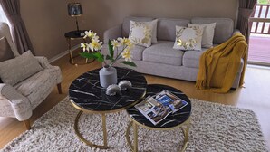 Een woonkamer met een grijze bank, een rond bijzettafeltje met een tijdschrift, een witte vaas met bloemen en een lijst met een afbeelding aan de muur.