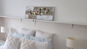 Een keurig opgemaakt slaapkamer met een bed, kussens, een nachtkastje met een lamp en een wandplank met een foto.