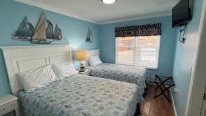 Une chambre avec deux lits, une table de chevet, une lampe, une fenêtre avec des stores et un téléviseur fixé au mur.