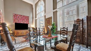 Un comedor con mesa de vidrio, sillas ornamentadas, candelabro y vistas de la ciudad a través de grandes ventanales.