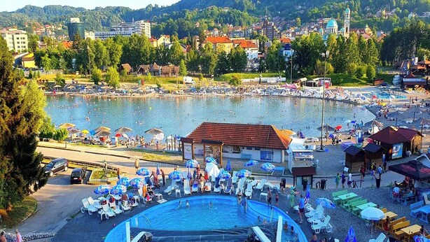 A swimming pool area with lounge chairs and umbrellas, surrounded by a lake and buildings in the background. - Apartment Meskovic Tuzla (Tuzla Grad)