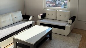 A modern living room with a white sofa, a coffee table, and a floor lamp. A window provides a view of buildings and greenery outside. - Apartment Meskovic Tuzla (Tuzla Grad)