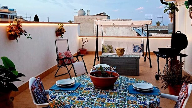 Eine Terrasse im Freien mit einem Tisch für zwei Personen, umgeben von weißen Stühlen, einem Grill und Blumentöpfen.