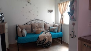 Ein Zimmer mit einem Bett, einer Wanduhr, einem Fenster mit Vorhängen und einem an der Wand befestigten Regal.