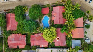 Ett flygbildsbild av ett semesterområde med flera byggnader med röda tak, en central pool och omgivande grönska, inklusive palmer.