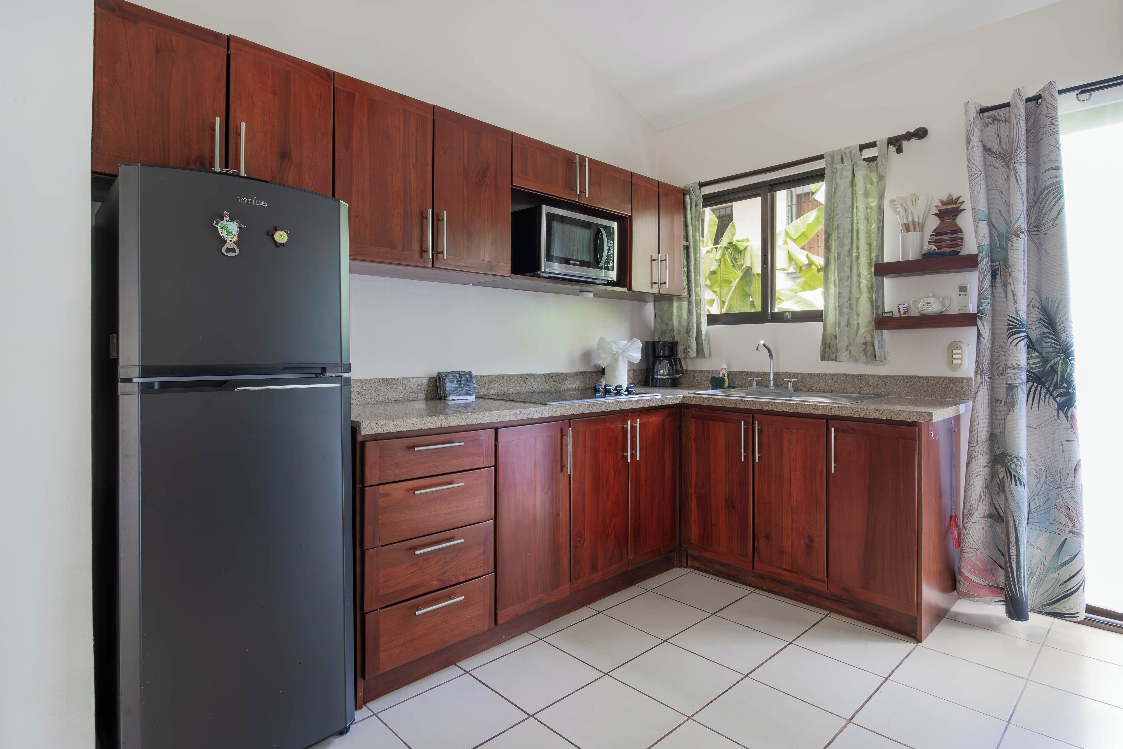 Una cocina con armarios de madera, un refrigerador negro, un microondas y una ventana con cortina.