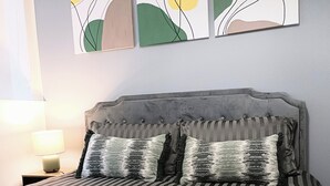 Una cama bien tendida con ropa de cama a rayas y dos almohadas, una lámpara de noche y arte abstracto en la pared sobre la cama.