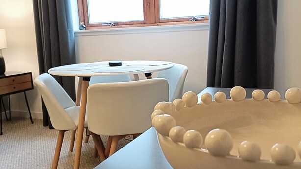 Una habitación con una mesa redonda, dos sillas, un bol grande con decoraciones esféricas, una lámpara y una ventana con cortinas.