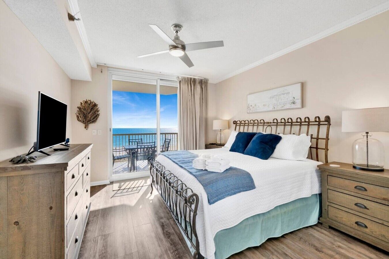 A hotel room with a bed, a TV, a desk, a chair, a balcony with a view of the sea, and a ceiling fan.