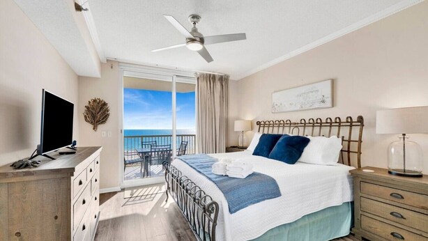 A hotel room with a bed, a TV, a desk, a chair, a balcony with a view of the sea, and a ceiling fan. - Beachfront Condo at Beach Club - Large Balcony! (Gulf Shores)