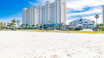 A beach with a volleyball net, palm trees, and a hotel with multiple floors and balconies.