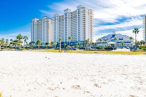 Ein Strand mit einem Volleyballnetz, Palmen und einem Hotel mit mehreren Etagen und Balkonen.