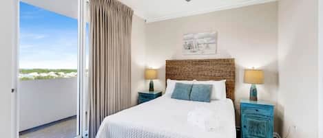 A bedroom with a bed, two bedside tables with lamps, a ceiling fan, a view through a sliding door, and a painting on the wall.